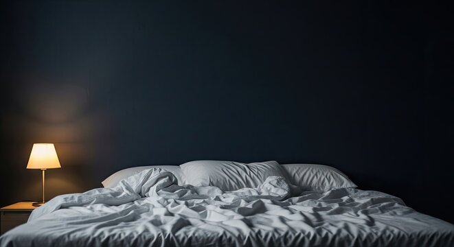 A messy bed with rumpled sheets and pillows in a dark room with a lamp on the nightstand.