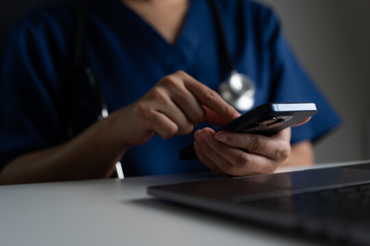 Doctor in scrubs holding and tapping on phone beside laptop, concept of remote medical service, digital diagnosis, mobile health applications, and healthcare technology in modern hospitals.