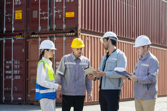 Logistics industrial workers in hard hats team discussing work plan at shipping container yard with safety gear, Construction and warehouse staff reviewing documents during outdoor briefing - Powered by Adobe