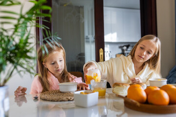 kids in bathrobes having breakfast together at home kitchen, girl helping sister pour juice