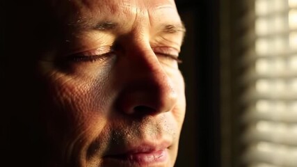 Close-up of a mans face with closed eyes, illuminated by sunlight filtering through blinds, creating a contemplative and peaceful mood. - Powered by Adobe