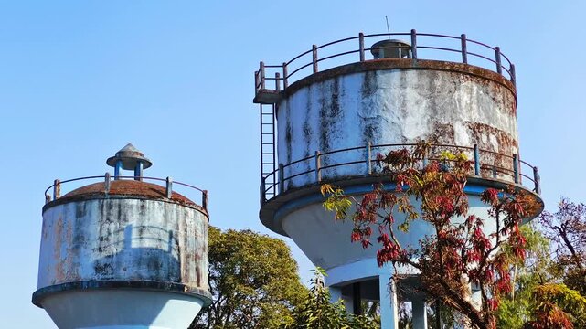 Two old water tanks rise above leafy trees under a clear sky, showing aging infrastructure and the industrial character of a small town environment.