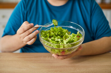 Obese man eating salad at home for healthy diet, weight loss and control. Chubby person holding fork with lettuce and cucumber to eat vegan food from glass bowl, enjoy low calorie meal and vitamins