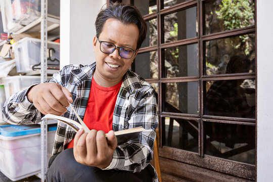 Indonesian southeast asian man dressed casually, looking relaxed and enjoying his leisure time while sitting outside and reading a book