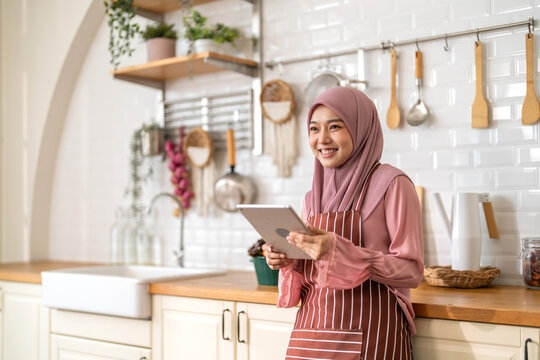 Smiling muslim woman wearing hijab use digital tablet in kitchen while preparing healthy food, online recipe search, home cooking and technology, halal cooking content, food blogger, healthy lifestyle