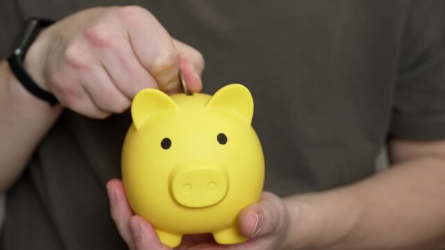 Caucasian hands dropping coin into yellow piggy bank, close-up of smartwatch wrist and casual shirt, steady insertion of spare change, scenes suggest student allowance, freelancer emergency