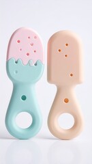 Fototapeta premium Two pastel-colored silicone ice cream-shaped teething toys, one pink and light teal, the other peach and light beige, featuring a handle with a ring, and swirls and dots