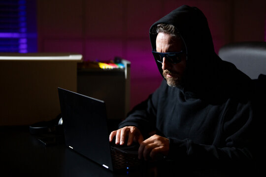 Hacker types on a laptop in a night office during a cyber attack. Hacker and stolen financial data concept. Digital money hacker. Cyber attack. Hacker in a hoodie using laptop in office.