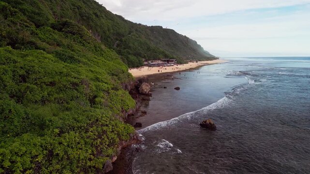 Drone video reveals a calm tropical beach along a wide Bali shoreline where low tide exposes shallow reefs beside a steep coastal green hill dotted with visitors enjoying the peaceful ocean atmosphere