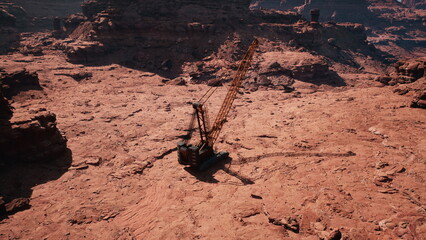 A large crane stands alone on the arid ground of a desolate desert. Surrounded by massive rock formations, it appears to be engaged in excavation or construction work under a clear blue sky.