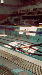 Bright sunlight filters through windows, casting playful shadows on the cool water of a swimming pool. Empty lanes wait for swimmers, inviting quiet moments of reflection.