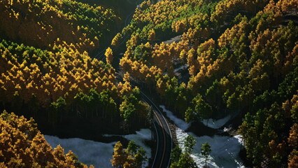 In a serene mountain valley, vibrant golden and green trees encircle a winding road. The sunlight casts warm hues, creating a peaceful autumn atmosphere. Snow remnants peek through the foliage.