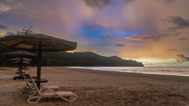 A tropical beach at sunset. A row of deck chairs under umbrellas on the sand. A calm ocean. The sky is illuminated by golden light near the horizon. Purple and pink clouds. Malaysia. Borneo.  - Powered by Adobe