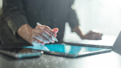 Close-up of a hand using a stylus on a digital tablet, showing a modern technology interface with data connections and analytics elements in a workspace setting.