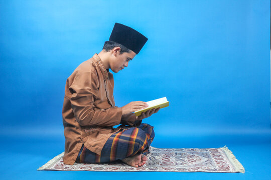 Muslim man reading a book sitting on a mat isolated on a blue background