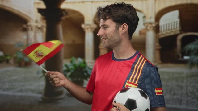 Man wearing red soccer jersey holding a black and white soccer ball and waving a small spanish flag in historic building courtyard with arches and plants; national pride celebration.