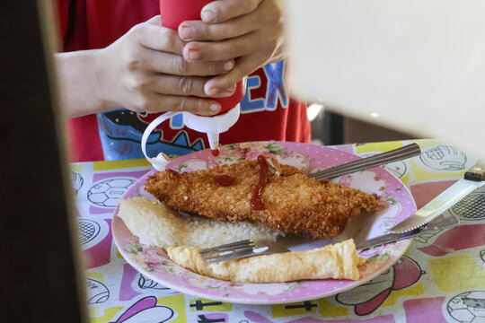 Child hand squeezing red ketchup from bottle onto golden fried fish fillet on bread. delicious crispy lunch meal ready for eating with fork and knife on plate