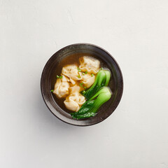 Chinese wonton soup with bok choy in black bowl on light background, view from above