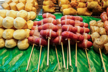 A vibrant selection of Thai street snacks on skewers, including bacon wrapped sausage, pork tofu meatballs, crab rolls, and fried dumplings, served on fresh banana leaves.