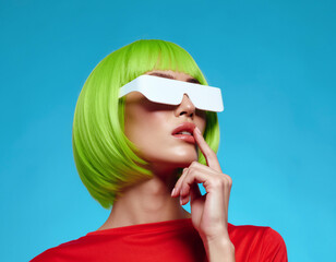 A woman with a vibrant green wig and white glasses poses against a blue backdrop.