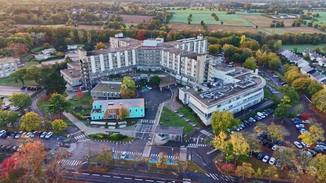 Drone establishing shot of CHU H&ocirc;pital Sud in Rennes, France, showing the sprawling hospital complex surrounded by tree-lined roads, parking areas, and green countryside on a sunny autumn morning.