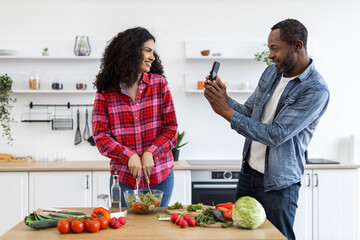 A couple is in the kitchen, with the woman preparing a salad while the man takes a photo with his phone.