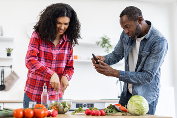 A couple is preparing a healthy meal together in a bright kitchen. The man is taking a photo of the woman with his phone.
