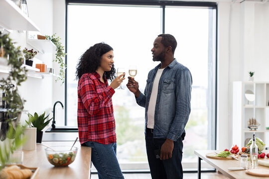 A diverse couple enjoys a moment together, toasting with wine glasses in a bright, contemporary kitchen setting.