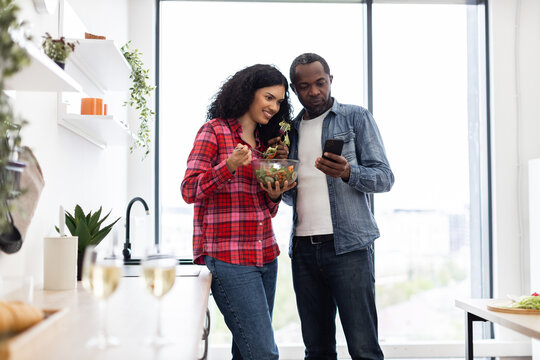 A diverse couple enjoys a healthy meal together in a modern kitchen, with one looking at a phone.