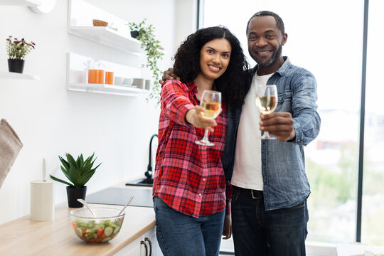 A smiling couple in a kitchen cheers with glasses of white wine, celebrating together.