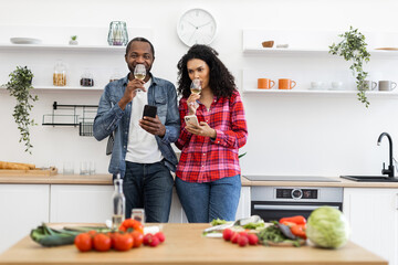 A diverse couple enjoys wine and uses their phones in a modern kitchen setting, surrounded by fresh produce.