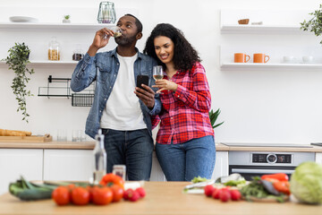 A couple is in their kitchen, looking at a phone while enjoying wine. Fresh vegetables are on the counter.