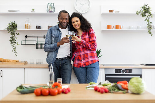 A smiling couple stands in a modern kitchen, toasting with glasses of wine, surrounded by fresh ingredients. - Powered by Adobe