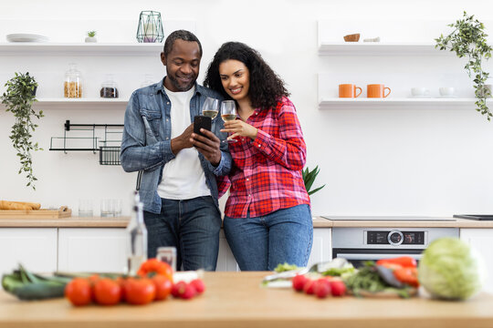 A diverse couple enjoys wine and looks at a phone in a modern kitchen with fresh produce.