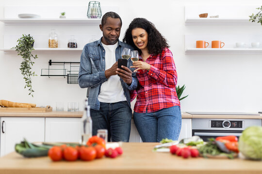 A diverse couple enjoys wine while looking at a smartphone in a modern kitchen, surrounded by fresh ingredients.