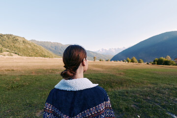 Naklejka premium Woman back view looking at expansive mountain valley landscape in nature outdoors, contemplative travel scene with distant peaks, grassy meadow and clear sky at golden hour.