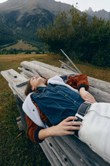 Naklejka premium woman bench mountains nature relaxed jacket denim outdoors lying on a wooden seat in a scenic meadow, casual fashion portrait showing leisure, travel and peaceful rest in countryside autumn