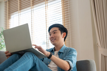 Young Asian man relaxing on comfortable couch, using laptop at home, happy boy chatting with friends in social network, spending weekend, watching movie, shopping online