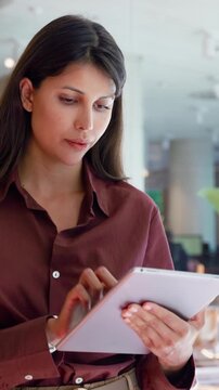 European businesswoman CEO using fintech tab application standing at workplace in office. Concentrated latin Hispanic mature professional business woman holding digital tablet for trading. Vertical
