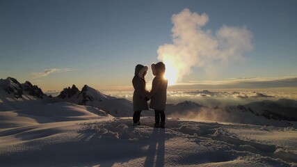 Two people in winter coats stand on a snowy mountain peak at sunrise, holding hands and facing each other with the sun casting long shadows and illuminating the snow-covered landscape around them