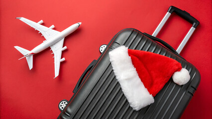 A black suitcase sits on a vibrant red background, topped with a Christmas hat. A small airplane model is nearby, hinting at upcoming holiday travel adventures in December.