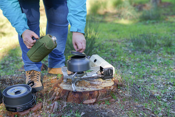 Person opening camping kettle on a portable stove, preparing hot drink while relaxing in tranquil forest during a morning outdoor adventure