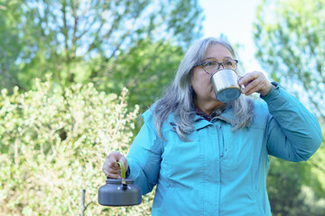 Senior woman relaxing outdoors, drinking from a metal mug, holding a camping kettle. Concept of peaceful outdoor living