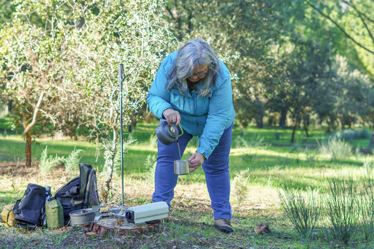 Senior woman pouring hot tea into a cup while relaxing during a camping trip in a forest enjoying nature - Powered by Adobe