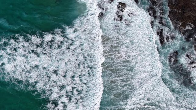 Aerial view of ocean waves crashing on rocky coastline, turquoise sea and white foam creating natural abstract pattern