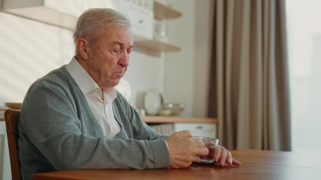 Lonely aged man feeling sad and drinking water in home kitchen, grandfather. Emotional male person missing and remembering life, widower thinking about past, healthcare in old age and aging of people