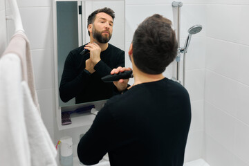 Man grooming beard in bathroom mirror while using electric trimmer during morning routine