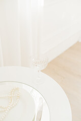 Elegant table setting with crystal glass, white plate, pearls, and silverware on a white table.
