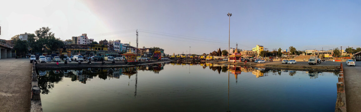 Panoramic view of a lakeside area in Nashik with buildings, ghats, parked vehicles, and calm water reflecting the entire cityscape during golden hour.