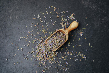 Dried lavender in wooden scoop on dark textured surface, scattered petals, herbal ingredient